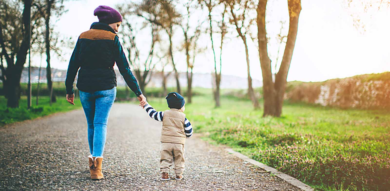 A mother and toddler walk side by side on a dirt path outside in the woods